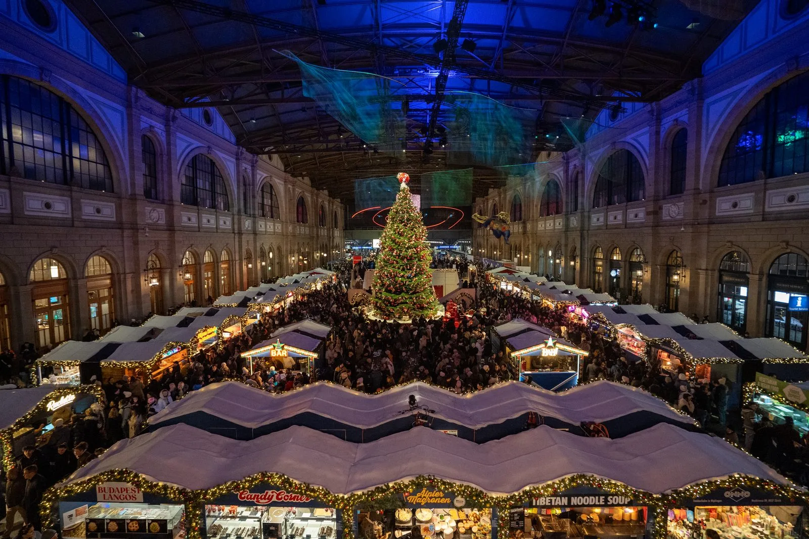 Weihnachtsmarkt Zürich Hauptbahnhof von oben: Markthäuschen, viele Besucher und ein Weihnachtsbaum unter dem blau beleuchteten Dach